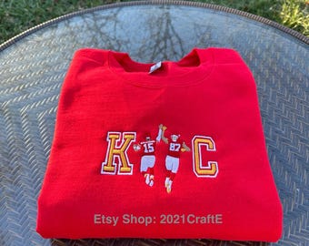 May include: Red sweatshirt with an embroidered design of the Kansas City Chiefs logo and two players in the team's uniform. The text "Etsy Shop: 2021CraftE" is printed on the bottom of the sweatshirt.