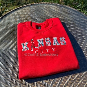 May include: A red sweatshirt with the words "KANSAS CITY" in white and teal lettering. A small illustration of a person playing basketball is on the left side of the text. The text "@Craft E Embroidery" is below the word "CITY".