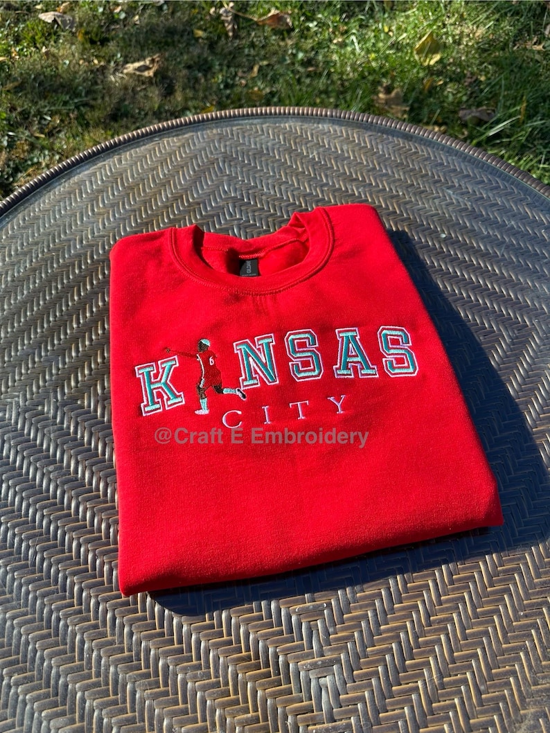 May include: A red sweatshirt with the words "KANSAS CITY" in white and teal lettering. A small illustration of a person playing basketball is on the left side of the text. The text "@Craft E Embroidery" is below the word "CITY".