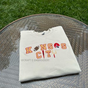 May include: A cream-coloured sweatshirt featuring embroidered text and graphics. The design includes the words "KANSAS CITY" in red and gold, a football, a helmet, and a goalpost. The text "@CRAFT E EMBROIDERY" is also visible.