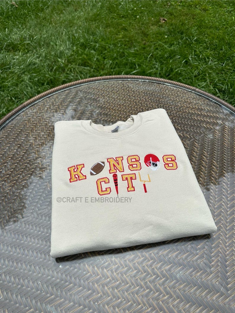 May include: A cream-coloured sweatshirt featuring embroidered text and graphics. The design includes the words "KANSAS CITY" in red and gold, a football, a helmet, and a goalpost. The text "@CRAFT E EMBROIDERY" is also visible.