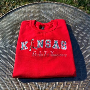May include: A folded red sweatshirt with the words "KANSAS CITY" in white and teal lettering. The sweatshirt has a small graphic of a person in a sports uniform. The text "@Craft E Embroidery" is below the city name.