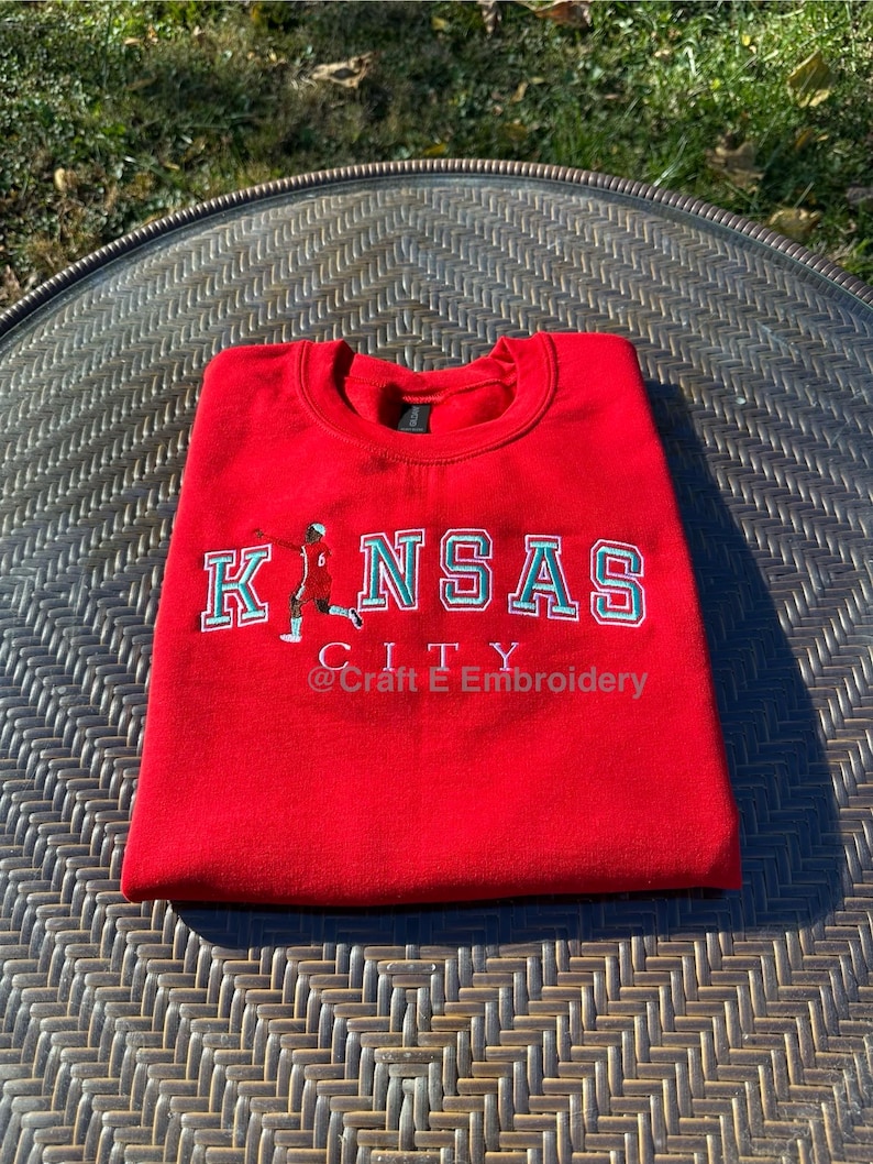 May include: A folded red sweatshirt with the words "KANSAS CITY" in white and teal lettering. The sweatshirt has a small graphic of a person in a sports uniform. The text "@Craft E Embroidery" is below the city name.