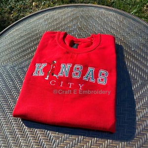 May include: A folded red sweatshirt with the words "KANSAS CITY" embroidered in white and teal. A small graphic of a person is on the left side of the text. The text "@ Craft E Embroidery" is below the text.