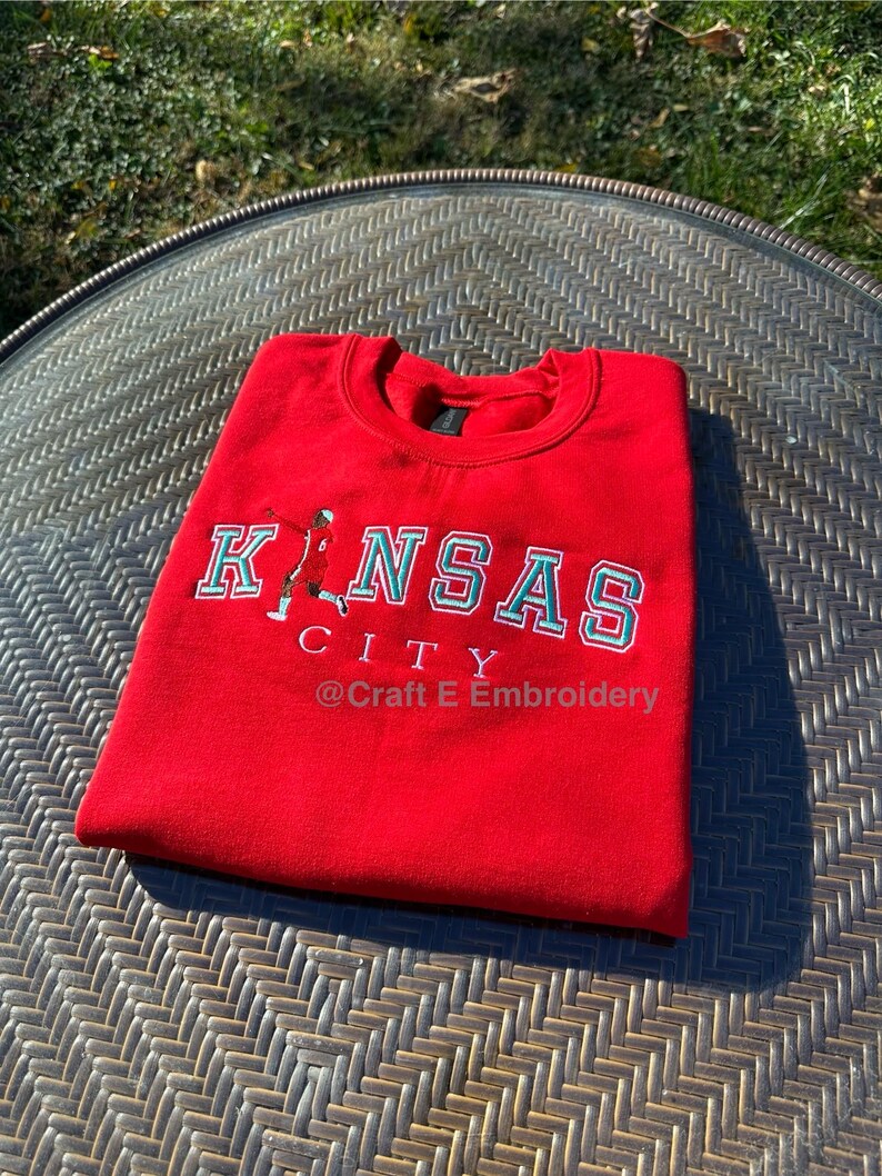 May include: A folded red sweatshirt with the words "KANSAS CITY" embroidered in white and teal. A small graphic of a person is on the left side of the text. The text "@ Craft E Embroidery" is below the text.