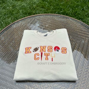 May include: A cream-coloured sweatshirt featuring the embroidered words "KANSAS CITY" in red and gold. The design includes a football, a helmet, and a goalpost. The text "@CRAFT E EMBROIDERY" is below the main text.