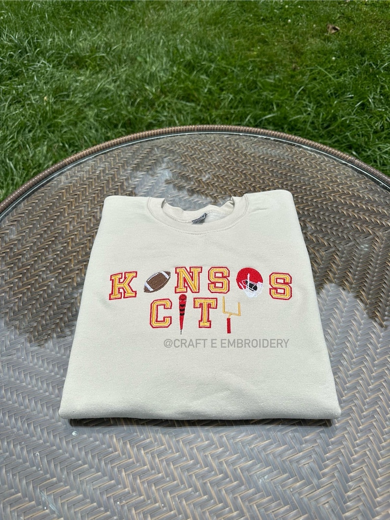 May include: A cream-coloured sweatshirt featuring the embroidered words "KANSAS CITY" in red and gold. The design includes a football, a helmet, and a goalpost. The text "@CRAFT E EMBROIDERY" is below the main text.