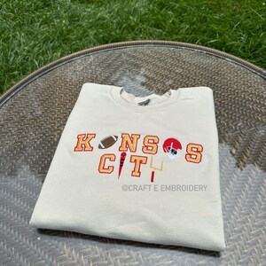 May include: A cream-coloured sweatshirt featuring the embroidered text "KANSAS CITY" in red and gold, accompanied by football imagery. The design includes a football, a helmet, and a goalpost. The text "@CRAFT E EMBROIDERY" is also visible.