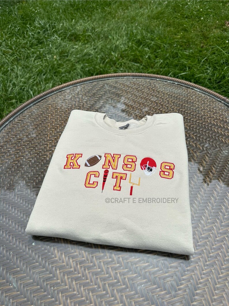 May include: A cream-coloured sweatshirt featuring the embroidered text "KANSAS CITY" in red and gold, accompanied by football imagery. The design includes a football, a helmet, and a goalpost. The text "@CRAFT E EMBROIDERY" is also visible.