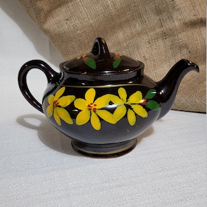 May include: A dark brown ceramic teapot with a lid, handle, and spout. The teapot is decorated with yellow floral designs and green leaves. The teapot sits on a small base and is suitable for serving tea.