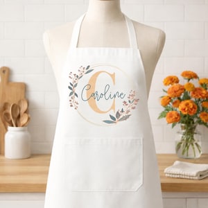 May include: White apron with a large front pocket, featuring a floral wreath design. The wreath surrounds the letter "C" and the name "Caroline" in a script font. The apron is displayed on a mannequin.