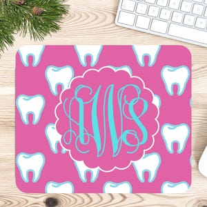 Puede incluir: Un mousepad rosa con un patrón de dientes blancos con detalles azules. Un marco festoneado blanco rodea un monograma turquesa. El mousepad es rectangular y está sobre una superficie de madera.