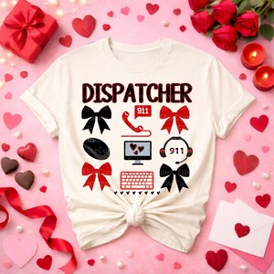 以下が含まれることがあります： 黒と赤で「DISPATCHER」の文字が入ったクリーム色のTシャツ。リボン、電話、コンピュータ画面、ヘッドセット、キーボードなど、さまざまなグラフィックが特徴です。背景にはハート、バラ、キャンドルが含まれています。