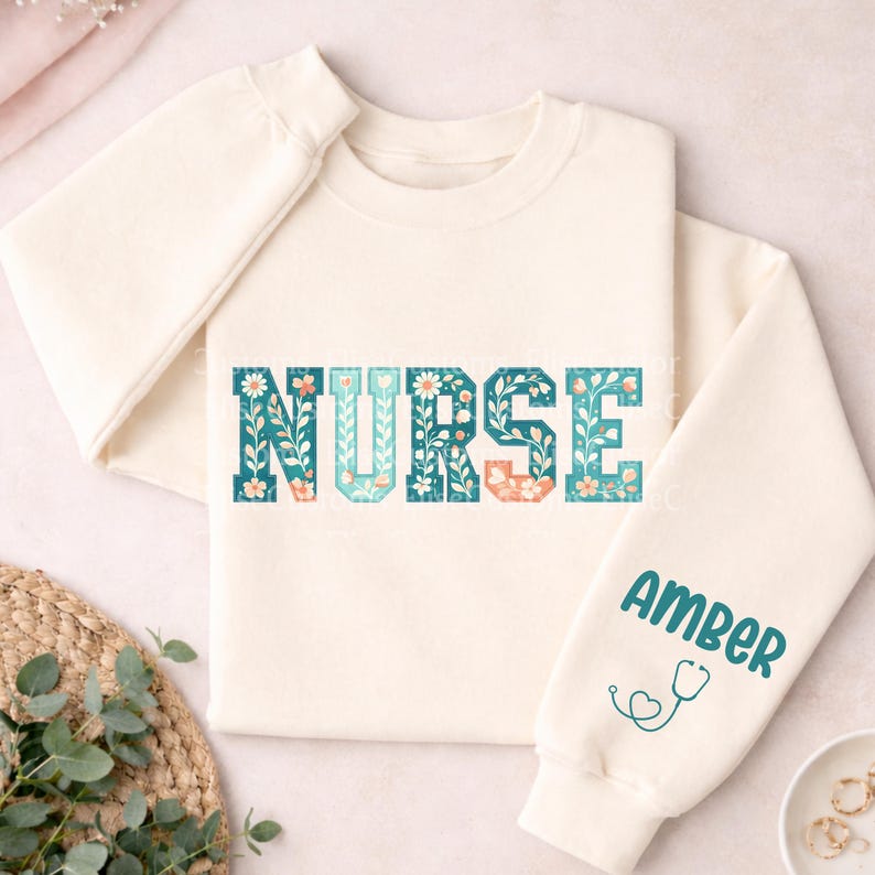 May include: Cream-colored sweatshirt with the word "NURSE" in a floral design across the chest. The letters are teal, blue, and peach. The right sleeve has the name "AMBER" in teal with a teal stethoscope graphic.
