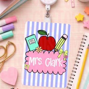 May include: A teacher's ID badge with a blue and white striped background. The badge features a red apple, a green notebook, and a yellow pencil. The name "Mrs. Clark" is written in pink script.