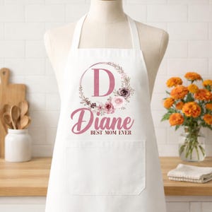 May include: White apron with a pocket, featuring a floral wreath design around the letter "D" and the name "Diane" in a cursive font. The text "BEST MOM EVER" is below the name. The apron is displayed on a mannequin.