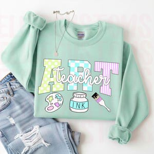 May include: A mint green sweatshirt with the words "ART Teacher" in a colorful design. The letter "A" has a polka dot pattern, the "R" has a checkered pattern, and the "T" has a striped pattern. The sweatshirt also features art-related graphics.