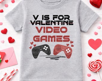 May include: A light gray t-shirt with the text "V IS FOR VALENTINE VIDEO GAMES" in black and red. Two game controllers are below the text, one black and one red, with small hearts. The shirt is surrounded by Valentine's Day decorations.