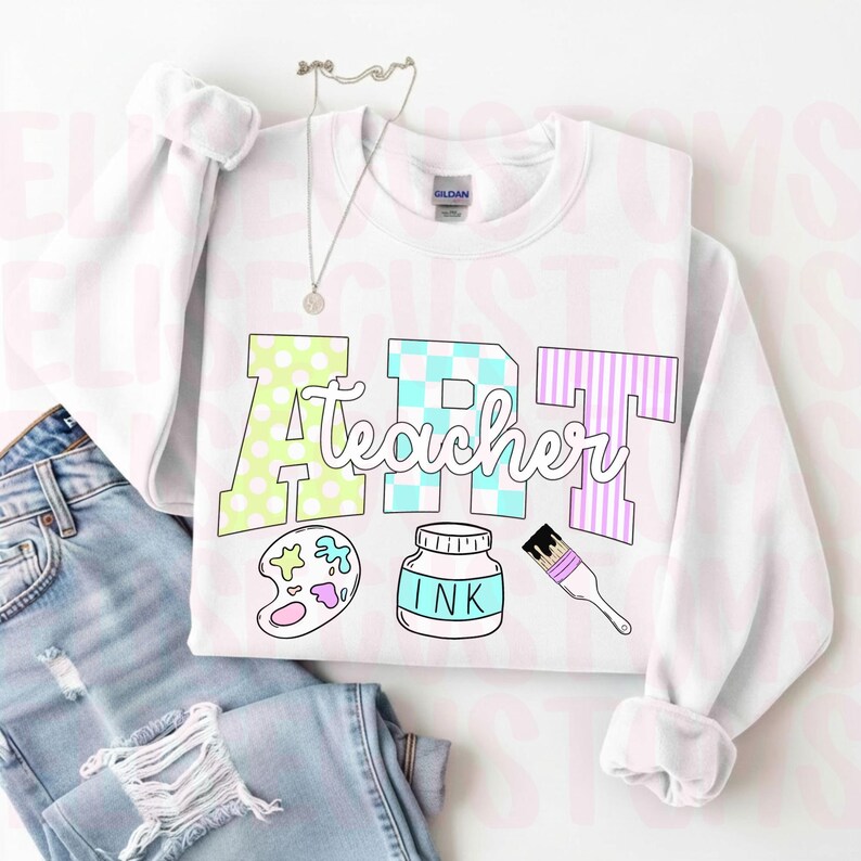 May include: White sweatshirt with the words "ART Teacher" in a colorful design. The letter "A" is light green with white polka dots, "R" is blue and white checkered, and "T" is purple and white striped. Below the text are art-related illustrations.