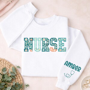 May include: White sweatshirt with the word "NURSE" in teal, peach, and white floral lettering. The right sleeve has the name "AMBER" in teal lettering, along with a teal stethoscope graphic. The sweatshirt is a crew neck style.