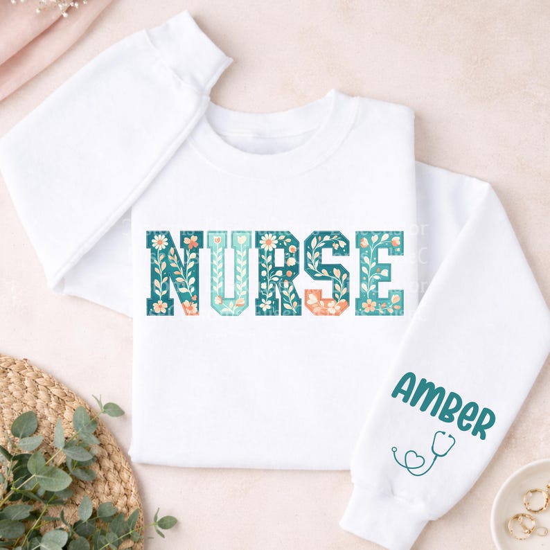 May include: White sweatshirt with the word "NURSE" in teal, peach, and white floral lettering. The right sleeve has the name "AMBER" in teal lettering, along with a teal stethoscope graphic. The sweatshirt is a crew neck style.