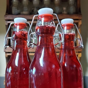 May include: Three glass bottles with red liquid and white lids, secured with metal clamps. The bottles are arranged in a row on a wooden surface. A single red rose is in the foreground.