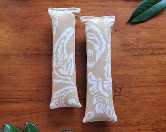 May include: Two beige catnip toys with a white floral pattern. The toys are made of fabric and are rectangular in shape.