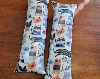 May include: Two white cat-themed fabric catnip toys. The toys are shaped like long rectangular pillows with a white background and a pattern of colorful cats with different expressions. The text "MILK" appears on both toys.