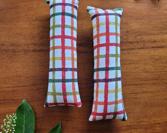 May include: Two long, rectangular catnip toys with a plaid pattern in shades of green, red, orange, and white. The toys are on a wooden surface.