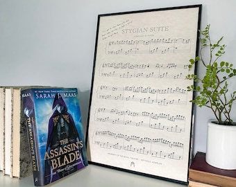 May include: A framed sheet music print with the title "Stygian Suite" composed by Pytor. The sheet music is property of the Royal Theater,  Bythold Adarlan. A book titled "The Assassin's Blade" by Sarah J. Maas is on a shelf below the print. A small potted plant is on a stack of books to the right of the print.