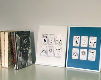 May include: Two framed prints of black and white illustrations of tarot cards. The prints are framed in white and blue frames. The prints are on a white surface with a book titled "Six of Crows" in the background.