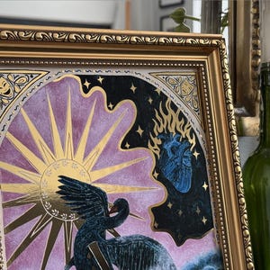 Op de afbeelding: Een ingelijst drukwerk met een gouden lijst en een paarse en zwarte achtergrond. De print toont een zwarte vogel met een gouden zon en een blauw hart.