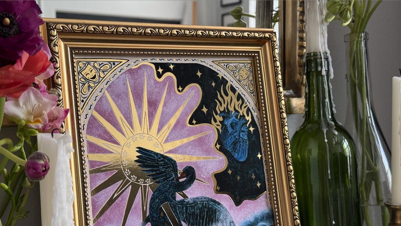 Op de afbeelding: Een ingelijst drukwerk met een gouden lijst en een paarse en zwarte achtergrond. De print toont een zwarte vogel met een gouden zon en een blauw hart.