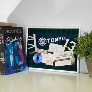Op de afbeelding: Een ingelijst prikbord met een groene en zwarte geruite achtergrond. Het bord is versierd met een blauw-witte vlag met de tekst "TOMMEN 13", een krantenknipsel uit "The Irish Times" en andere ephemera.