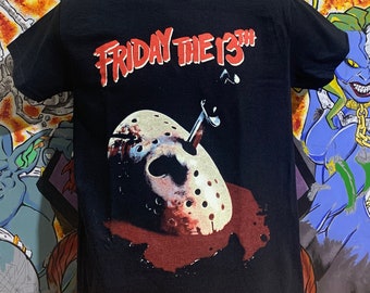 May include: Black t-shirt featuring the text "Friday the 13th" in red, above an image of a hockey mask with a knife. The shirt is displayed on a black mannequin head against a colorful graffiti background.