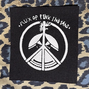 May include: A black square patch with white text and a peace symbol design. The text reads "flux of pink indians." The patch is set against a leopard print background in shades of tan, blue, and black.