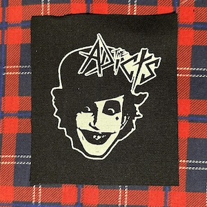 May include: Black square patch featuring a white illustration of a smiling face with the text "The Adicts" above the head. The patch is on a red, blue, and white plaid fabric background.