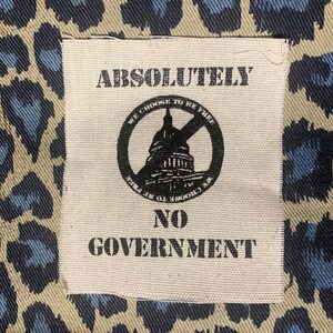 May include: A white patch with black text that reads "ABSOLUTELY NO GOVERNMENT" with a black and white graphic of the US Capitol building with a circle and slash through it. The text "WE CHOOSE TO BE FREE" is written around the graphic. The patch is on a brown and blue leopard print fabric.
