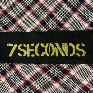 May include: Black rectangular patch with the text "7 SECONDS" in yellow stencil font, with a crosshair symbol. The patch is set against a plaid pattern in black, white, and red.