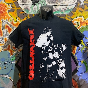 May include: Black t-shirt with a white graphic print of a punk rock band, Discharge, performing on stage. The band's name is printed in red on the side of the shirt.
