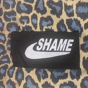 May include: A black patch with the word "SHAME" in white, stylized like a popular brand logo, is sewn onto a fabric with a tan, black, and blue leopard print pattern. The fabric has a repeating animal print.
