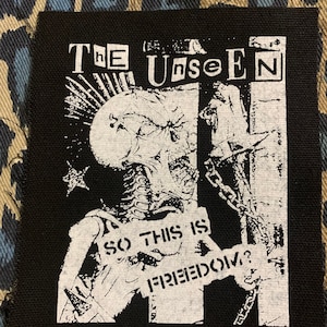 May include: Black and white patch with the text "The Unseen" and "So this is freedom?"  The patch features a skeleton-like figure with a chain and a skull.