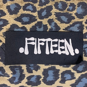 May include: Black fabric patch with white text that reads ".FIFTEEN."