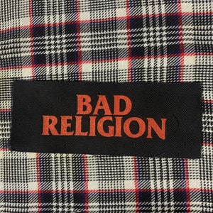 May include: A black rectangular patch with the words "BAD RELIGION" in orange lettering is centered on a plaid fabric. The plaid pattern features a mix of white, black, and red lines, creating a classic checkered design.