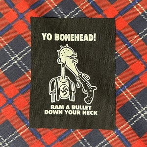 May include: Black and white iron-on patch with a cartoon character holding a gun and the text "Yo Bonehead! Ram a bullet down your neck."