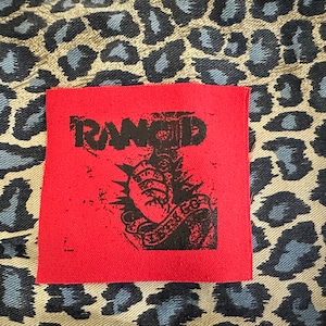 May include: Red fabric patch with a black and white graphic of a hand holding a heart with a barbed wire design and the word "RAMMED" above it.