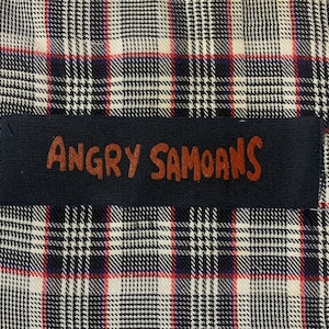 May include: A black and white plaid fabric with a black label that says "Angry Samoans" in brown lettering.