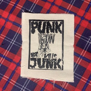 May include: A rectangular patch with the words "PUNK HEROIN NOT KILLS JUNK" in black block letters. The patch features a skeletal figure and is set against a beige background. The background is a red and blue plaid pattern.