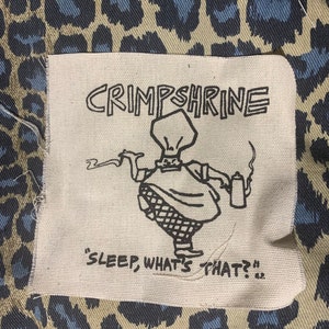 May include: A white fabric patch with black text and a cartoon illustration. The text reads "Crimpshrine" and "Sleep, what's that?" The illustration depicts a person wearing a chef's hat and holding a teapot.