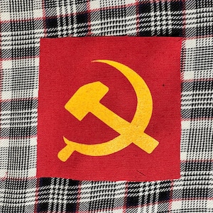May include: A red fabric patch with a yellow hammer and sickle symbol. The patch is sewn onto a black, white, and red plaid fabric.